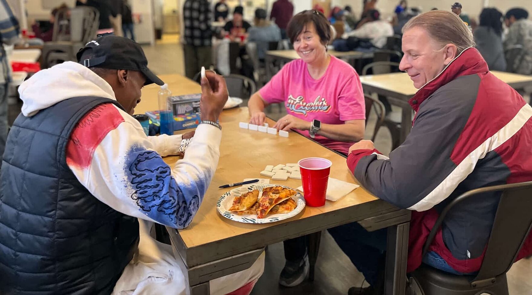 team leaders Jennifer (in a pink inExcelsis t-shirt), and Mark( inExcelsis bus driver in a grey-red jacket) are playing domino with a guest at ComFe.