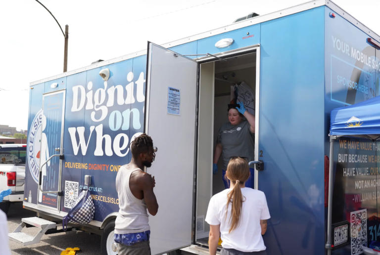 Two volunteers sanitizing a shower stall with a guest holding the door ready to go in.